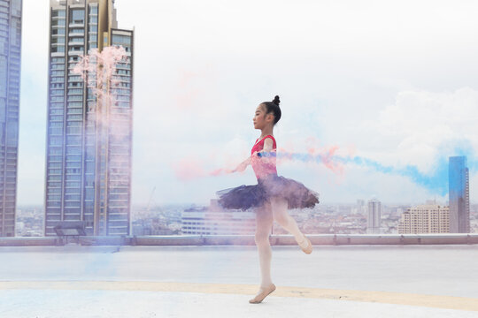 Asian ballerina ballet girl dancing with colour smoke on rooftop and skyscraper city view background. Female ballet dancer practicing dancing. Young ballerina ballet girl dancing