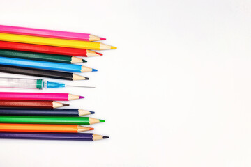 An empty syringe with a needle lies among colored pencils