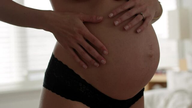 Pregnant woman at 28 weeks rubs bare belly in front of mirror in morning sunlight - medium shot