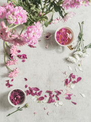 Fototapeta na wymiar Cup with pink flowers tea on light desk with peonies flowers bunch and petals . Top view. Frame