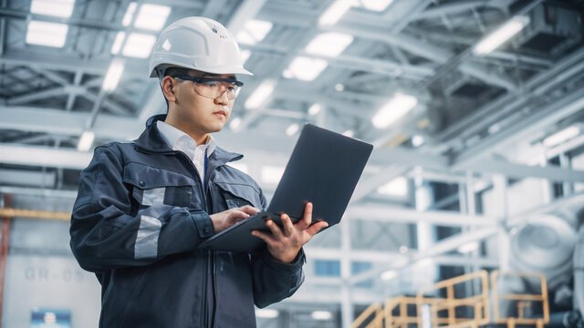 Portrait of a Professional Heavy Industry Asian Engineer/Worker Wearing Safety Uniform and Hard Hat Uses Laptop Computer. Confident Chinese Industrial Specialist Standing in a Factory Facility.