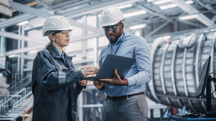 Two Diverse Professional Heavy Industry Engineers Wearing Safety Uniform and Hard Hats Working on Laptop Computer. African American Technician and Female Worker Talking on a Meeting in a Factory.