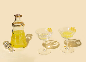 One unique and antique bohemian decanter and two luxurious crystal glasses with yellow cocktails and lemon slice on it against beige background.