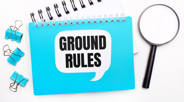 Office Table With Blue Notepad, Paper Clips, Magnifying Glass And White Card With Text GROUND RULES. Top View, Flat Lay. Stylish Workplace