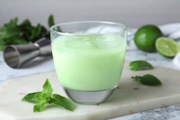 Delicious mint liqueur with ice cubes and green leaves on white marble table, closeup