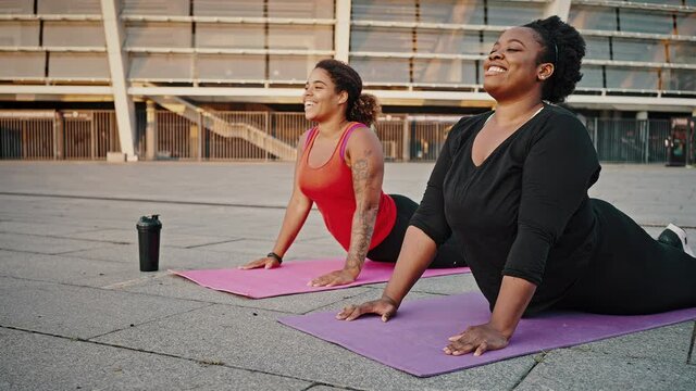Two flexible overweight african american ladies practicing yoga training, stretching bodies on fitness mats in city area