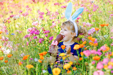 Portriat of adorable, charming toddler girl with Easter bunny ears eating chocolate bunny figure in flowers meadow. Smiling happy baby child on sunny day with colorful flowers, outdoors.