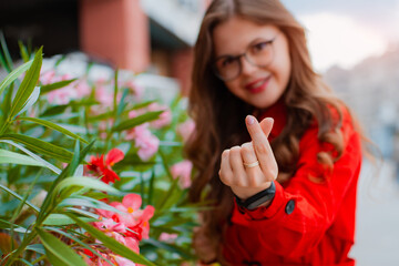 Positive funky girl in glasses making korean love sign or heart gesture while standing outside