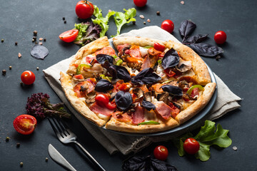 Savory Homemade Meat Pizza with Pepperoni, Bacon, tomatoes and basil on a dark background.