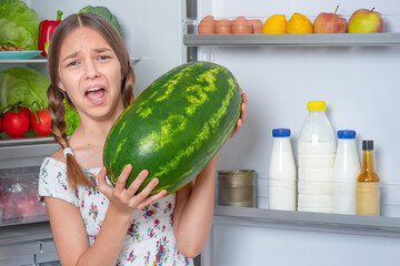 Fototapeta premium Beautiful young teen girl holding big Watermelon while standing near open fridge in kitchen at home
