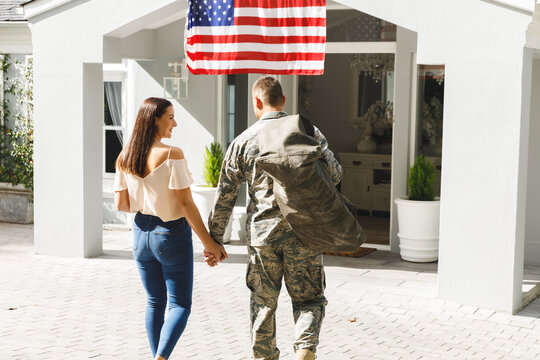 Caucasian male soldier holding hands with wife outside house decorated with american flag
