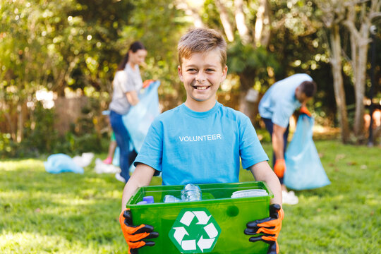 Portrait of smiling caucasian son holding recycling box, cleaning up countryside with parents
