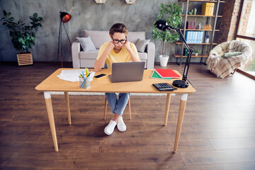 Photo of tired upset young lady wear yellow t-shirt sitting table arms cheekbones working modern gadget indoors room home