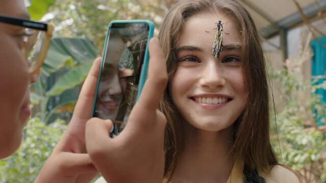 beautiful butterfly on girls face with happy friend taking photo using smartphone friends having fun in zoo wildlife sanctuary sharing nature excursion on social media 4k footage