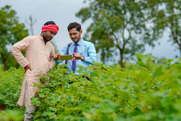 Young indian agronomist or banker showing some information to farmer in smartphone at agriculture field.