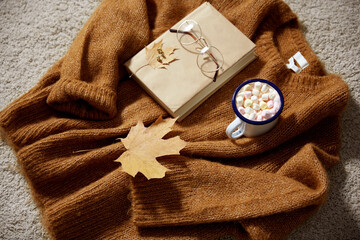 Fototapeta na wymiar season, leisure and objects concept - camp mug full of marshmallow, book, glasses and autumn leaves on sweater