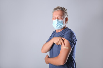 Senior man in protective mask pointing at arm with bandage after vaccination on grey background....