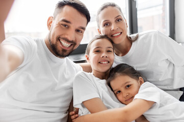 Fototapeta premium family and people concept - happy mother, father and two daughters taking selfie in bed at home