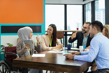 Asian Muslim business woman in hijab headscarf sitting on wheelchair presenting of her work to corporate colleagues in meeting in the modern office. diverse corporate colleagues and multicultural