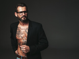 Portrait of handsome confident stylish hipster lambersexual model. Sexy modern man dressed in black jacket. Naked torso with tattoos.Fashion male posing in studio on dark background. In spectacles - obrazy, fototapety, plakaty