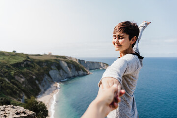 Fototapeta na wymiar happy Girlfriend holding hand of boyfriend during hiking on top on the cliff. Sea view. Relax and nature concept