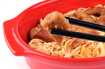 Morceau de porc au caramel tenu avec des baguettes chinoises et une plat de nouilles chinoises au second plan