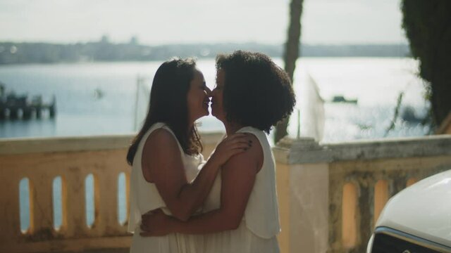 Medium shot of African-American lesbians hugging before wedding. Side view of smiling brides kissing on sunny day on embankment. LGBT wedding, love, romantic relationship concept