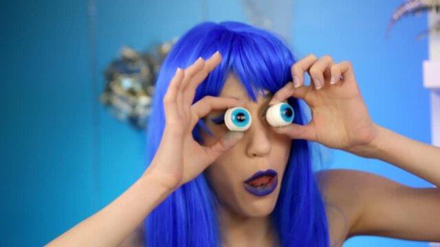 A woman in a blue wig measures her toy eyes and grimaces at the camera.