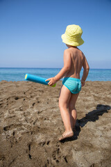 Boy stands on the beach, holding a water gun toy and looking towards the sea