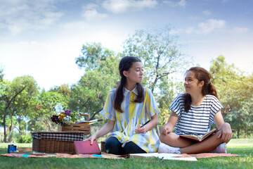 Two pre-teen girls sit on picnic mat and look at each other at park, summer holidays