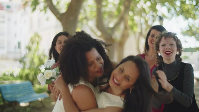 Beautiful lesbians celebrating wedding with friends in park. Smiling girls in evening wears enjoying happy moments, applauding, congratulating and laughing together. LGBT wedding, party concept