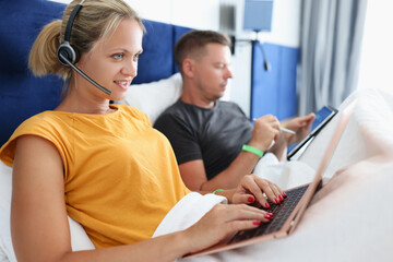Woman in headphones lying in bed and working at laptop next to man with tablet