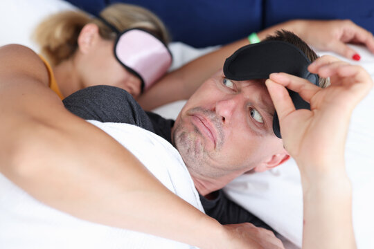 Surprised man lifting his sleep mask and looking at woman hugging him in bedroom