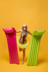 full length of happy woman in sunglasses and swimsuit holding inflatable mattresses and standing with swim ring on yellow.