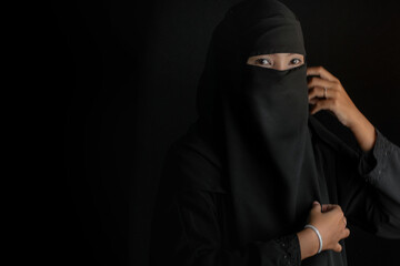 Beautiful Arabian woman in black hijab, scarf and black veil, woman portrait wearing Nigab in black background, looking at camera