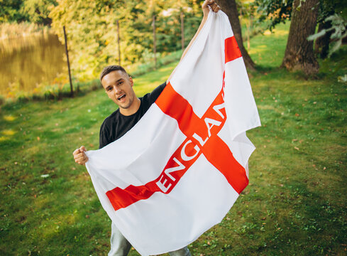 Portrait of young handsome man holding flag of England isolated over natural park landscape