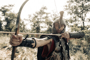 Ancient warrior archer aims and shoots from bow among meadow. - obrazy, fototapety, plakaty