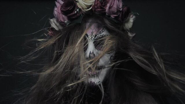 Halloween makeup, a girl in the image of a cute and good death Santa Muerte or a sugar Mexican skull calmly looks at the camera with hair in the wind in slow motion.