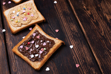 Toast with chocolate paste and peanut butter sprinkled with colorful hearts on a dark wooden background.
