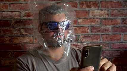 A plastic bag is worn on the head. The man has dark glasses. Looks at a smartphone. Halloween holiday. Party. Ecology problems. Businessman. Close-up.