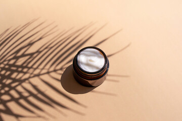 Cosmetic white creme in dark glass jar on beige background with palm leaves shadow. Beauty product skin care package.