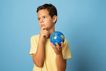 Pensive school boy holding a world globe isolated over blue background