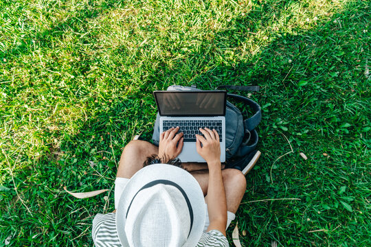 Top view of an unrecognizable young white digital nomad working remotely on a laptop sitting on a lawn
