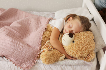 a little girl in a blonde T-shirt is sleeping at home under a pink blanket hugging a teddy bear