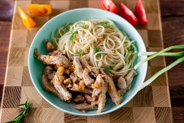 Fried meat and spaghetti on a plate on a wooden background with tomatoes and herbs.