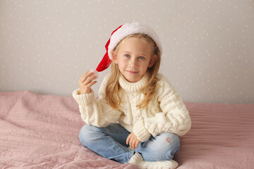 a beautiful little blonde girl is sitting at home on the bed in a winter sweater and a Santa claus hat new year Christmas