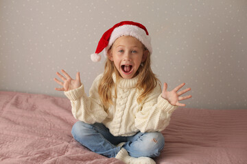 a beautiful little blonde girl is sitting at home on the bed in a winter sweater and a Santa claus hat new year Christmas