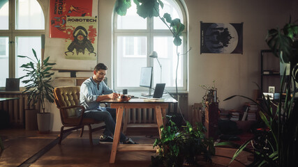Young Handsome Man Works on a Desktop Computer in Creative Agency in Authentic Loft Office. Renovated Stylish Design with House Plants, Artistic Posters and Big Rounded Windows.