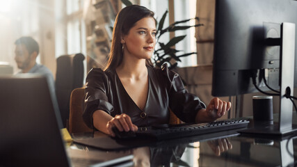 Portrait Shot of a Stylish Employee Working on Computers in Creative Agency in Loft Office. Beautiful Female Manager Typing Emails. Sunny Renovated Space with Artistic Posters and Big Windows.