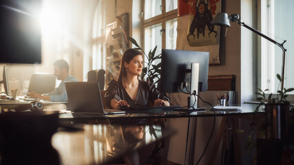 Two Stylish Employees Working on Computers in Creative Agency in Loft Office. Beautiful Manager Typing Correspondence Emails. Sunny Renovated Space with Plants, Artistic Posters and Big Windows.